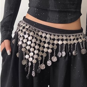 Pode incluir: Cinto prateado com detalhes de moedas penduradas. O cinto &eacute; usado sobre uma pe&ccedil;a de roupa preta. As moedas s&atilde;o circulares e presas a uma corrente. O cinto foi projetado para ser usado na cintura.