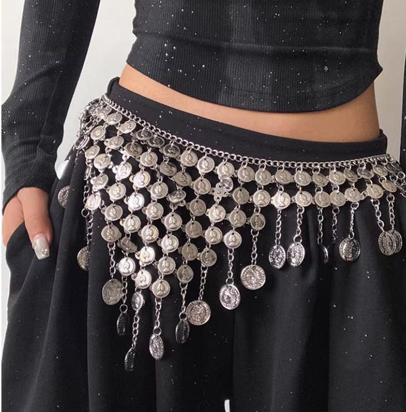 Pode incluir: Cinto prateado com detalhes de moedas penduradas. O cinto &eacute; usado sobre uma pe&ccedil;a de roupa preta. As moedas s&atilde;o circulares e presas a uma corrente. O cinto foi projetado para ser usado na cintura.