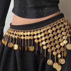 Pode incluir: Um cinto de dan&ccedil;a do ventre dourado com detalhes de moedas e correntes. O cinto &eacute; usado sobre uma saia preta e apresenta fileiras de moedas circulares com correntes e moedas penduradas. &Eacute; um acess&oacute;rio decorativo.