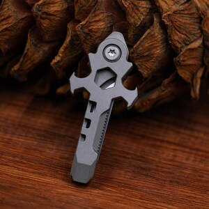 May include: A grey multi-tool featuring a knife, hex wrench, and screwdriver. The tool has a distinctive, cross-shaped design with a textured grip and a small, star-shaped screw. The tool is made of metal and is designed for everyday carry.
