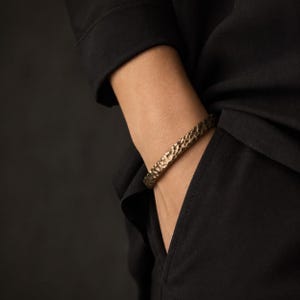 Peut inclure: Un bracelet manchette en bronze texturé. Le bracelet a un aspect martelé et est porté au poignet. Le fond est de couleur noire unie. Le bracelet est un bijou simple mais élégant.
