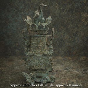 May include: A bronze-colored, ornate vessel with a bird-like figure atop a crown-like structure. The vessel is approximately 3.9 inches tall and weighs about 7.8 ounces. The body of the vessel features intricate carvings and handles.