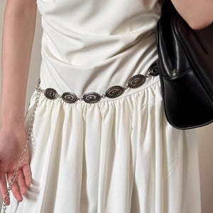 Pode incluir: Um cinto de metal prateado com enfeites ovais e um detalhe de corrente. O cinto &eacute; usado sobre um vestido branco. Uma bolsa preta &eacute; segurada na imagem.