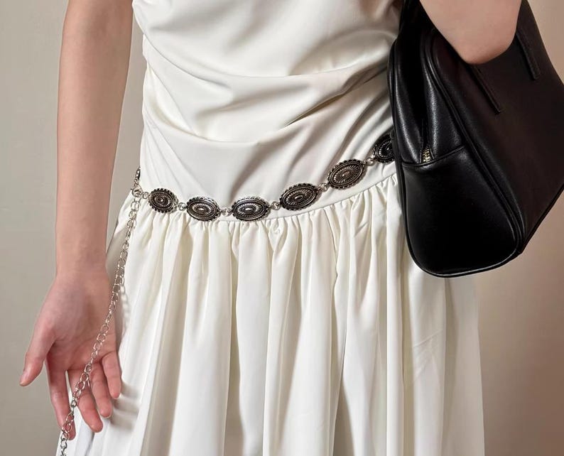 Pode incluir: Um cinto de metal prateado com enfeites ovais e um detalhe de corrente. O cinto &eacute; usado sobre um vestido branco. Uma bolsa preta &eacute; segurada na imagem.