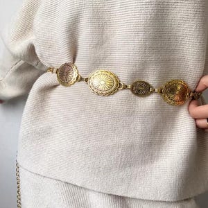 Pode incluir: Um cinto de metal dourado com medalh&otilde;es decorativos. O cinto apresenta v&aacute;rias formas e padr&otilde;es em relevo. O cinto &eacute; usado sobre um su&eacute;ter creme, criando um acess&oacute;rio elegante.