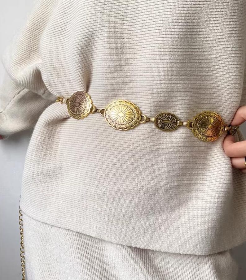 Pode incluir: Um cinto de metal dourado com medalh&otilde;es decorativos. O cinto apresenta v&aacute;rias formas e padr&otilde;es em relevo. O cinto &eacute; usado sobre um su&eacute;ter creme, criando um acess&oacute;rio elegante.