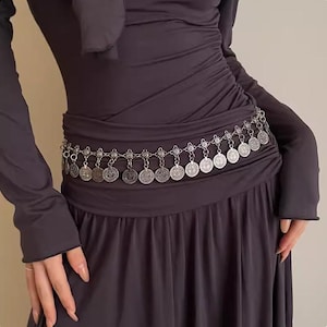 Pode incluir: Um cinto prateado com pingentes de moedas. O cinto &eacute; usado sobre um vestido cinza escuro. As moedas circulares t&ecirc;m padr&otilde;es detalhados. O design do cinto adiciona um elemento decorativo ao traje.