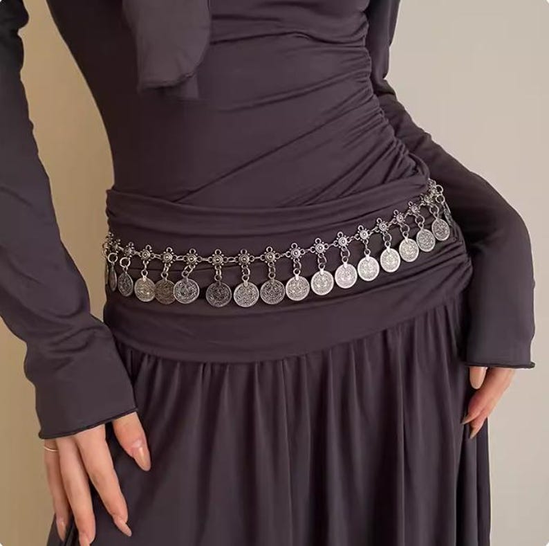 Pode incluir: Um cinto prateado com pingentes de moedas. O cinto &eacute; usado sobre um vestido cinza escuro. As moedas circulares t&ecirc;m padr&otilde;es detalhados. O design do cinto adiciona um elemento decorativo ao traje.