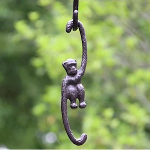 Peut inclure: Une figurine de singe en métal foncé suspendue à un crochet incurvé. Le singe est détaillé avec de la fourrure visible et s'accroche à une barre incurvée. L'arrière-plan est vert flou, suggérant un cadre extérieur.