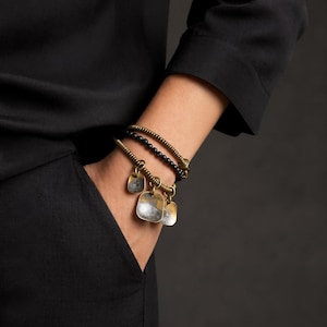 Può includere: Un set di tre braccialetti. Uno è un braccialetto in ottone a spirale con ciondoli quadrati, un altro è un braccialetto di perline con perline nere e il terzo è un braccialetto a spirale in ottone. I braccialetti sono indossati su un polso.