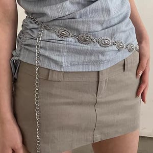 Pode incluir: Cinto de corrente prateada com elementos decorativos ovais. Usado sobre um top azul claro e uma saia c&aacute;qui. A corrente pende, adicionando um detalhe elegante. O design do cinto &eacute; intrincado e atraente, adequado para v&aacute;rias roupas.