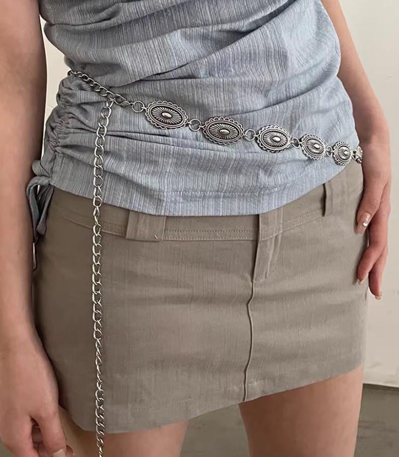 Pode incluir: Cinto de corrente prateada com elementos decorativos ovais. Usado sobre um top azul claro e uma saia c&aacute;qui. A corrente pende, adicionando um detalhe elegante. O design do cinto &eacute; intrincado e atraente, adequado para v&aacute;rias roupas.