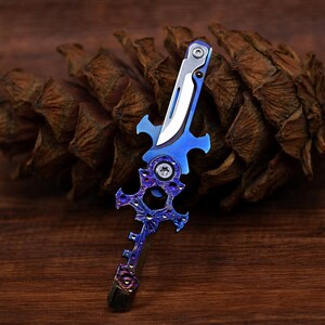 May include: A decorative knife with a blue and silver blade and handle. The handle features an ornate, stylized design with purple accents. The knife is set against a brown, textured background, suggesting a natural setting.
