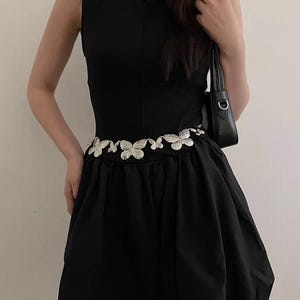 Pode incluir: Vestido preto sem mangas com cinto de borboletas prateadas. O vestido tem um corpete justo e uma saia rodada. O cinto &eacute; o ponto focal, adicionando um toque decorativo. O estilo geral &eacute; elegante e moderno.