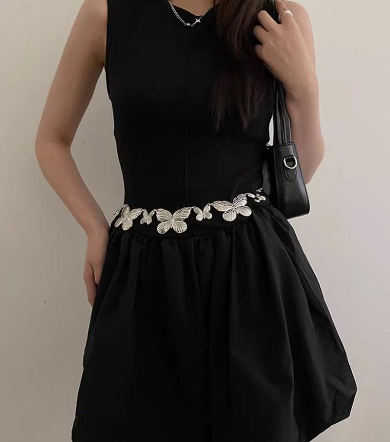 Pode incluir: Vestido preto sem mangas com cinto de borboletas prateadas. O vestido tem um corpete justo e uma saia rodada. O cinto &eacute; o ponto focal, adicionando um toque decorativo. O estilo geral &eacute; elegante e moderno.