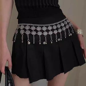 Pode incluir: Corrente de cintura em metal prateado com detalhes florais e contas penduradas. A corrente &eacute; usada sobre uma saia plissada preta. &Eacute; um acess&oacute;rio decorativo.