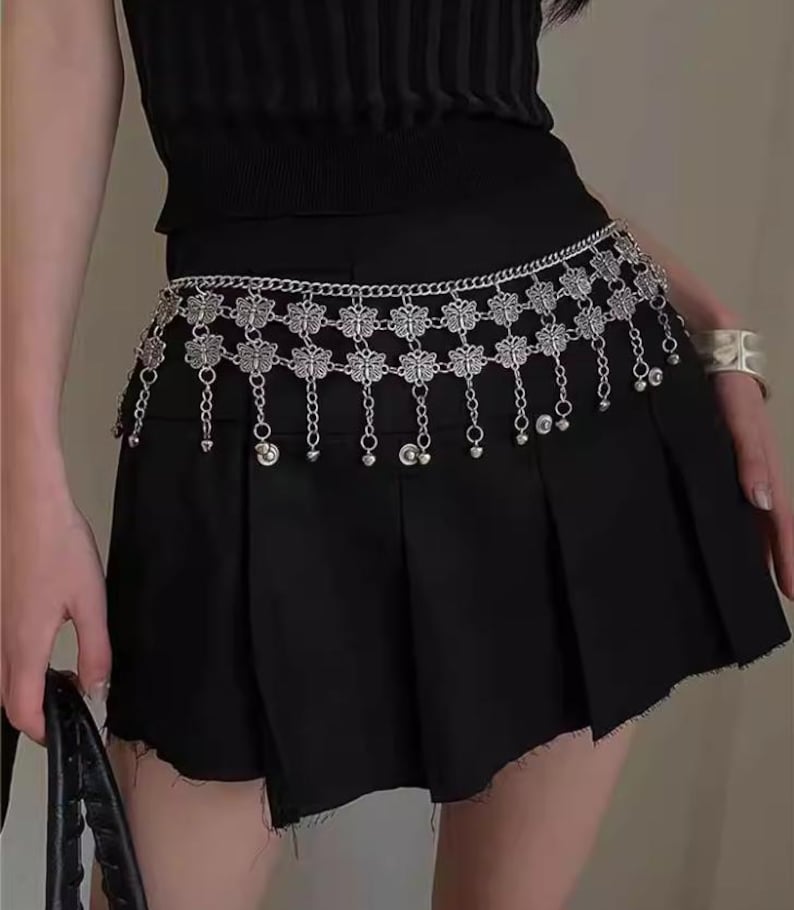 Pode incluir: Corrente de cintura em metal prateado com detalhes florais e contas penduradas. A corrente &eacute; usada sobre uma saia plissada preta. &Eacute; um acess&oacute;rio decorativo.