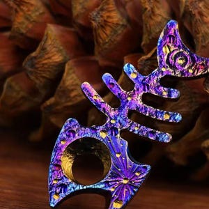 Könnte beinhalten: Ein schillernder, fischgrätenförmiger Ring. Der Ring zeigt ein Farbspektrum aus Blau-, Lila- und Goldtönen mit strukturierter Oberfläche. Das Design umfasst eine kreisförmige Öffnung für den Finger und wirkt künstlerisch und einzigartig.