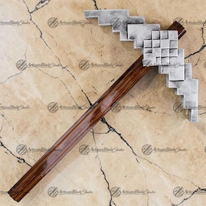 Puede incluir: Un pico con una cabeza pixelada plateada y un mango de madera marrón oscuro. El pico está diseñado en un estilo retro y en bloques, que recuerda a un videojuego. El mango mide aproximadamente 61 cm de largo.
