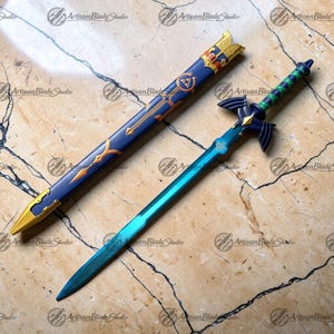 Puede incluir: Una espada azul y dorada con una vaina a juego. La espada tiene una hoja turquesa y una empuñadura detallada con detalles verdes. La vaina es azul con detalles dorados y una punta dorada. El texto "Artisan Blade Studio" es visible.