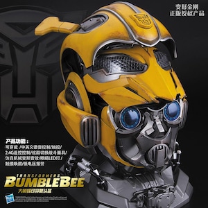 Może przedstawiać: Żółto-srebrny hełm Bumblebee Transformers z wykończeniem typu weathered, o mechanicznym designie, z niebieskimi oczami LED i wysuwaną maską. Hełm zawiera chiński tekst opisujący funkcje, takie jak sterowanie głosowe i efekty dźwiękowe.