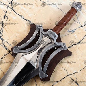 Può includere: Immagine dettagliata di una spada con una lama in acciaio lucido e un'impugnatura avvolta in pelle marrone. La spada presenta una guardia decorativa color argento ed è montata su una placca di legno scuro. Il testo "Artisan Blade Studio" è visibile.