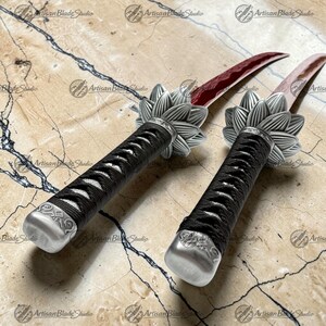 May include: Two daggers with curved, deep red blades and black cord-wrapped handles. Each handle features a silver-toned metal pommel and a silver-toned metal guard shaped like a multi-petaled flower. The blades have a visible wavy temper line.