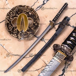 May include: A Japanese katana sword with a black handle and scabbard. The blade is silver with a patterned design. The sword is displayed with a decorative, circular guard with an ornate, gold-colored design. The words "Artisan Blade Studio" are visible.