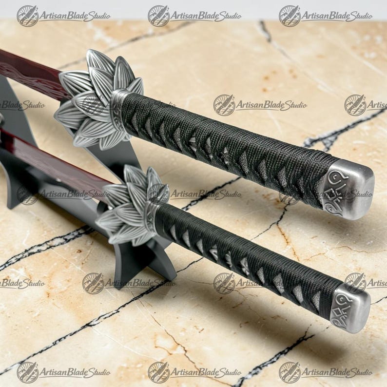 May include: Two swords with dark red blades and black cord-wrapped handles. Each handle features a silver-colored metal tsuba shaped like a lotus flower and a matching silver-colored pommel with engraved detailing, resting on a black stand.