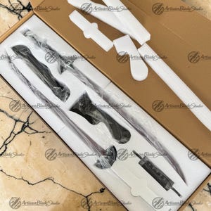 May include: A long, slender black sword with a curved blade and a detailed hilt, encased in clear plastic within a custom-fitted white foam insert inside a brown cardboard box. The set includes a separate small black component in the foam.