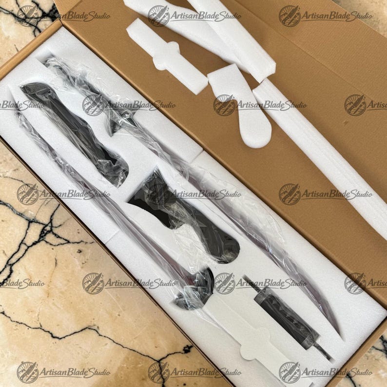 May include: A long, slender black sword with a curved blade and a detailed hilt, encased in clear plastic within a custom-fitted white foam insert inside a brown cardboard box. The set includes a separate small black component in the foam.