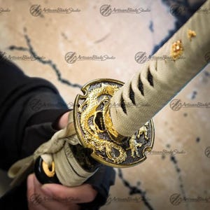 Könnte beinhalten: Nahaufnahme eines Katana-Schwertes. Der Griff hat ein goldenes Drachendesign auf schwarzem Hintergrund. Der Griff ist mit einem hellbeigen Stoff umwickelt. Die Klinge ist nicht sichtbar. Das Schwert wird in einer Hand gehalten.