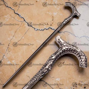 May include: A silver walking cane with a detailed handle and a long, slender shaft. The handle has a textured pattern. The cane is displayed on a marble surface. The text "Artisan Blade Studio" is visible. The cane's length is approximately 90 cm.