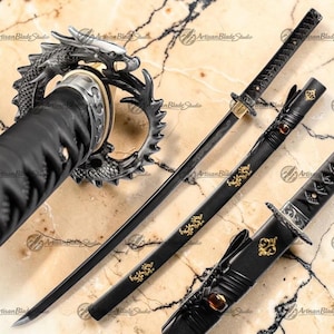 May include: A black katana sword with a dragon-shaped tsuba and gold accents. The scabbard is black with gold dragon designs. The blade is visible and has a slight curve. The handle is wrapped in black material. The text "Artisan Blade Studio" is visible.