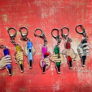 May include: A collection of seven keychains featuring colorful microphone designs held by illustrated hands. Each keychain has a silver-colored ring and clasp. The microphones are various colors, including blue, yellow, pink, green, red, and black.