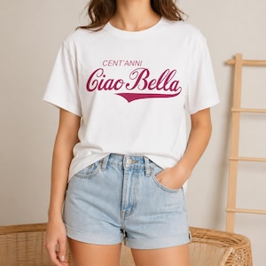 以下が含まれることがあります： 白いTシャツに、バーガンディ色の筆記体で「CENT'ANNI Ciao Bella」と書かれています。半袖で、ライトブルーのデニムショーツと合わせています。モデルは明るい色の壁の前に立っています。