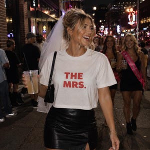 May include: A white short-sleeved crop top with the text THE MRS. printed in red letters, paired with a black leather mini skirt featuring a small front slit, a white bridal veil, and a black shoulder bag, worn in a nighttime urban setting.