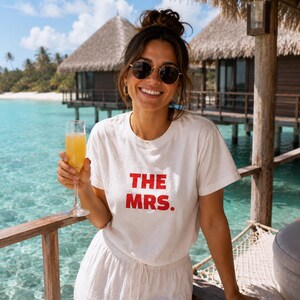 May include: A white short-sleeved t-shirt featuring the text THE MRS. printed in bold red capital letters on the front. The shirt has a crew neckline and a relaxed fit, paired with white drawstring trousers, round sunglasses, and gold hoop earrings.