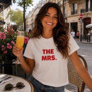 May include: A white short-sleeved t-shirt with the text THE MRS. printed in bold red capital letters on the front. The shirt features a crew neckline and a straight hem, paired with blue denim shorts. The design is simple and casual in style.