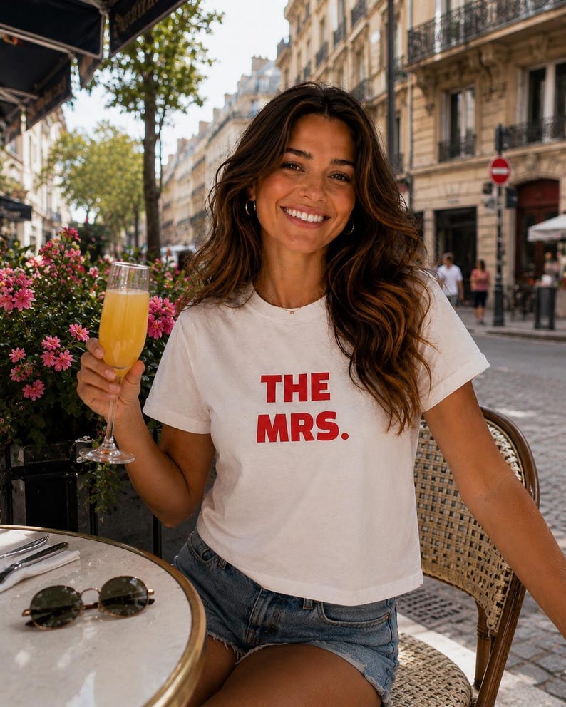 May include: A white short-sleeved t-shirt with the text THE MRS. printed in bold red capital letters on the front. The shirt features a crew neckline and a straight hem, paired with blue denim shorts. The design is simple and casual in style.