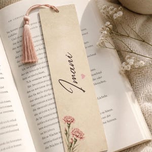 Könnte beinhalten: Ein rechteckiges Lesezeichen aus Papier mit cremefarbenem Hintergrund, das den Namen Imani in schwarzer Schreibschrift, ein kleines rosa Herz und eine florale Illustration von zwei rosa Nelken unten zeigt, mit einer rosa Quaste.