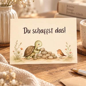 May include: A rectangular greeting card featuring a light cream background with the German text "Du schaffst das!" printed in dark brown. The illustration shows a green turtle stacking grey stones next to a small brown bird among flowers.