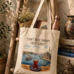 Könnte beinhalten: Eine beigefarbene Stofftasche mit dem aufgedruckten Text "I DON'T NEED THERAPY, I NEED TURKEY." Die Grafik zeigt eine Ansicht einer Uferpromenade mit einer Moschee, einem Boot und einem Teeglas auf einer Untertasse im Vordergrund.