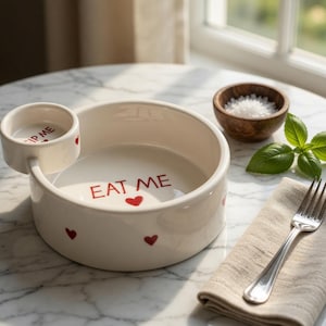 Può includere: Una ciotola da portata in ceramica bianca con una piccola ciotola per salse attaccata. Le parole "EAT ME" sono in rosso, insieme ad accenti a forma di cuore rosso. La ciotola per salse dice "DIP ME". Sono presenti anche una piccola ciotola di legno con sale, basilico e una forchetta.