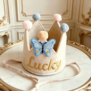 Peut inclure: Une couronne en feutre beige avec un papillon bleu appliqué et le mot "Lucky" brodé en or. La couronne est ornée de pompons aux couleurs pastel et possède une fine sangle élastique.