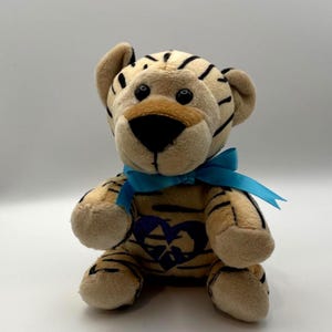 May include: A plush tiger toy with a tan body, black stripes, and a blue ribbon tied around its neck. The tiger has a heart design on its chest and black eyes. The toy is sitting upright.