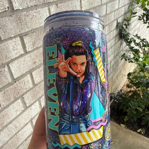 May include: A clear glass tumbler with a lid, filled with purple glitter. The tumbler features a colorful graphic of the character Eleven from the show Stranger Things, with the word "ELEVEN" in teal. The design includes geometric shapes and stars.