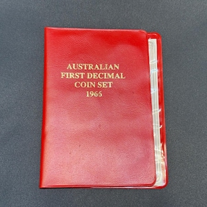 May include: A red coin album with gold lettering that reads "AUSTRALIAN FIRST DECIMAL COIN SET 1965". The album is closed and appears to be in good condition. The background is a dark grey surface.