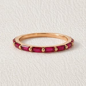 May include: A rose gold ring with a band of rectangular, deep red gemstones. Small, round gold accents are placed between each gemstone. The ring is set against a white background.