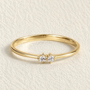 May include: A slender gold ring with two small, clear gemstones set close together. The ring has a thin band and a polished finish. The gemstones are round and sparkle. The ring is displayed on a neutral, textured surface.